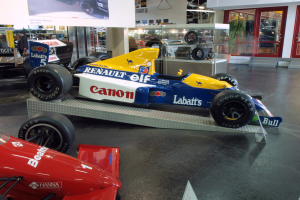 Eine Museumausstellung mit verschiedenen Rennwagen, darunter ein prominent ausgestelltes Renault F1-Auto, mit Besuchern, die beobachten, und informativem Text an einer Wand im Hintergrund.