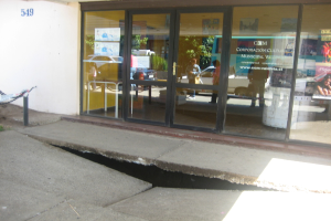 Eine gebrochene Oberfläche mit einer Glastür, einer Wand und einem Pfahl im Hintergrund, die sich bewegende Fahrzeuge und einige stehende Personen spiegeln.