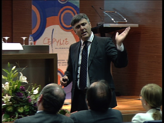 Ein Mann in einem schwarzen Blazer steht an einem Podium mit Mikrofonen, spricht in ein Mikrofon und hält einen Blumentopf oder einen Blumenstrauß, mit einem Tisch mit Gegenständen wie einem Buch und einer Wasserflasche daneben und drei Personen, die davor sitzen, vor einer Wand in Braun und einer bunten Tafel mit Text.