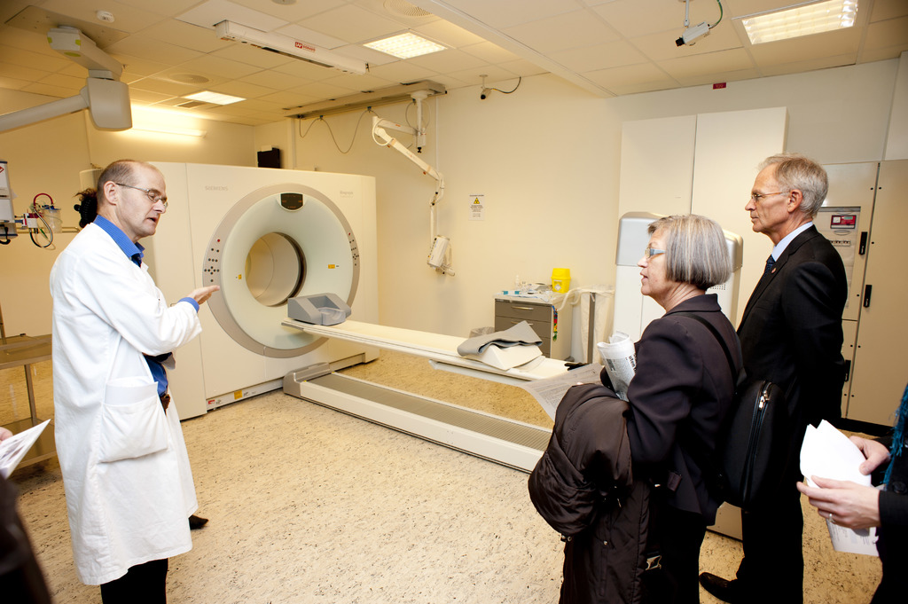 Ein Arzt in weißer Uniform erklärt eine CT-Scan-Maschine zwei Menschen in schwarzen Mänteln, die aufmerksam zuhören.