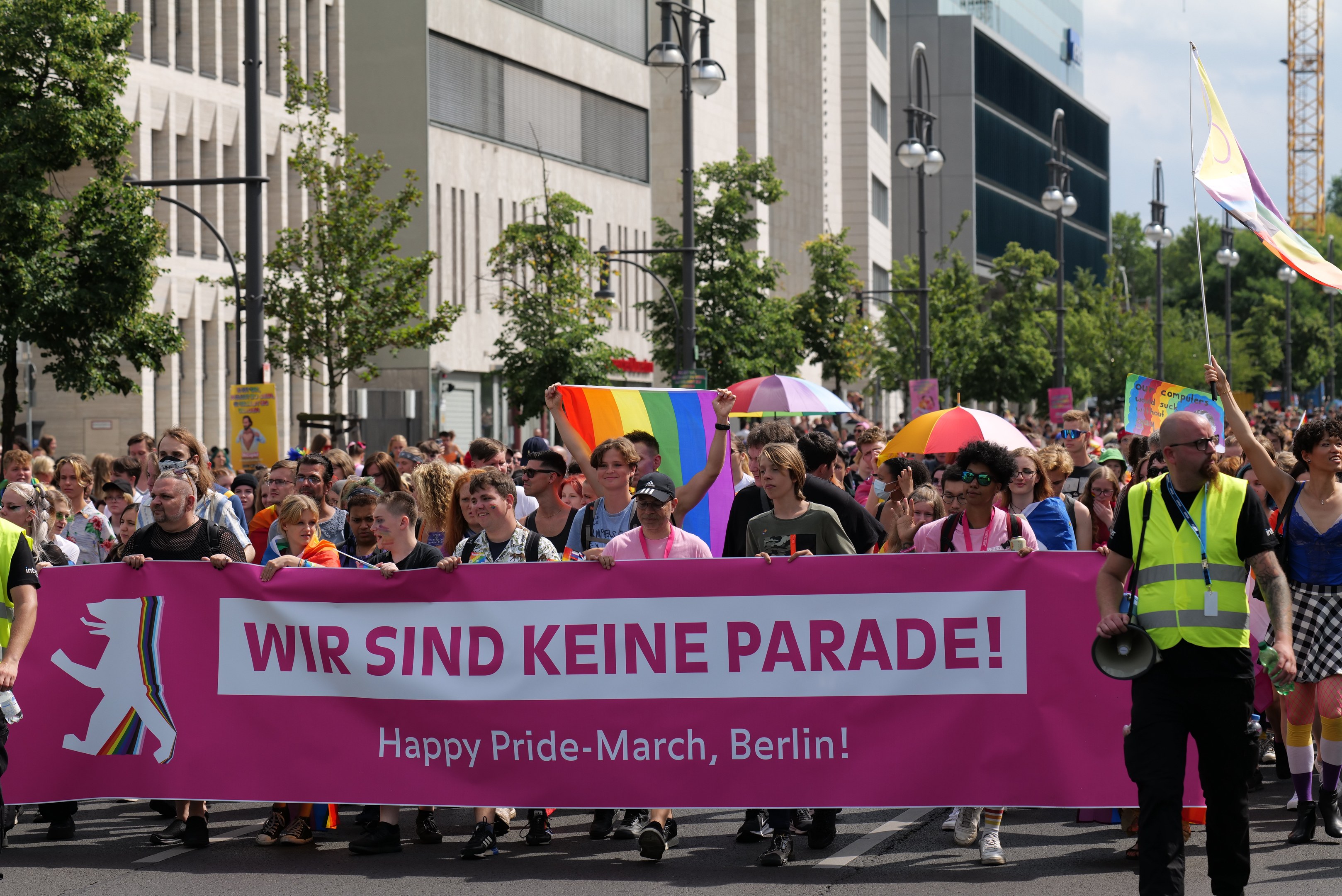 Eine Gruppe von Menschen marschiert bei einer Pride-Veranstaltung in Berlin, hält einen pinken Banner mit der Aufschrift "Happy Pride March Berlin" und trägt Schirme und Fahnen.