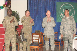 Vier Personen stehen, drei klatschen, eine sitzt auf einem Stuhl neben einer Pflanze, mit einem Banner im Hintergrund, alle tragen Militäruniform, mit Stühlen und einem Tisch im Sichtfeld.