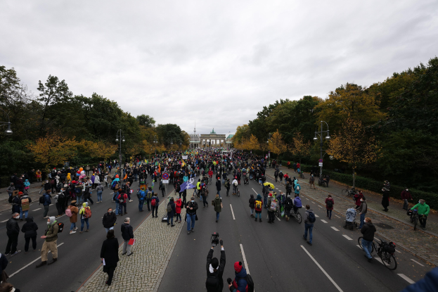 Eine große Gruppe von Menschen, die eine straßenbepflanzte Straße in Berlin entlangmarschieren, einige mit Kameras, mit einem Gebäude und einem klaren Himmel im Hintergrund.