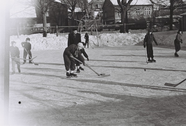 Eine Gruppe von Menschen, die Hockey auf einem Eisplatz umgeben von Schnee, Bäumen, Gebäuden mit Fenstern, Treppen mit Geländern und einem Lastwagen in der Umgebung spielen, dargestellt in Schwarz-Weiß.