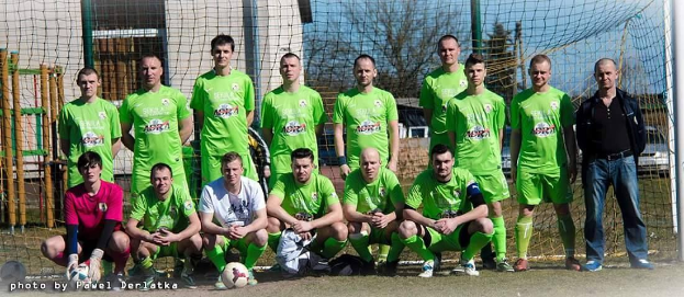 Eine Gruppe von Männern in grünen T-Shirts steht auf einem Fußballfeld in der Nähe eines Torpfostens, einige hocken, mit einem Ball davor und Gebäuden, Bäumen und Fahrzeugen im Hintergrund.
