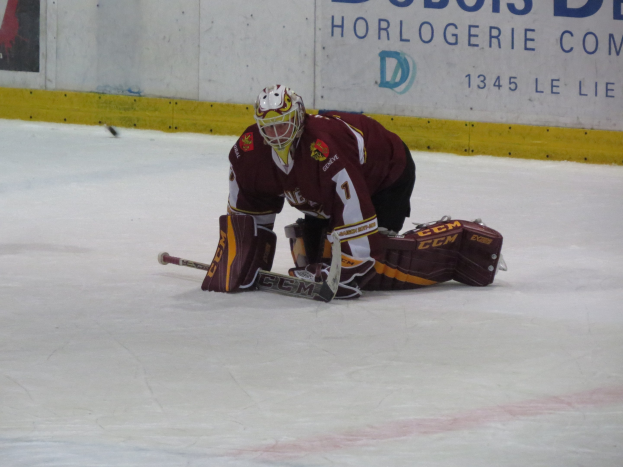 Eishockeyspieler in rot-gelber Uniform mit Helm, Handschuhen und Knieschonern beim Abwehren eines Schusses auf dem Eis, mit einer Wand mit Text im Hintergrund.