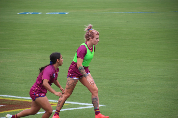 Zwei Frauen spielen Fußball auf einem Feld, eine trägt ein grünes T-Shirt und Shorts, die andere ein pinkes Kleid, beide tragen Sportkleidung und zeigen sichtbare Tattoos an Händen und Füßen.