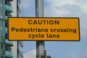 Ein gelbes Warnschild an einem Pfahl vor einem Gebäude, mit einem klaren blauen Himmel im Hintergrund, auf dem steht: "Füßgänger überqueren Fahrradweg."