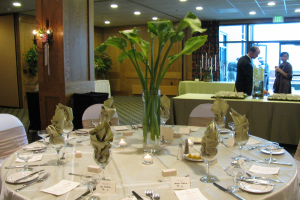 Ein Tisch mit einer weißen Decke, Tellern, Besteck, einem Glas und Baumzweigen, umgeben von Stühlen, mit einem grünen Tisch dahinter; ein Mann in einem schwarzen Blazer und ein Mädchen neben ihm, mit einer Tür im Hintergrund und zwei Männern dahinter.