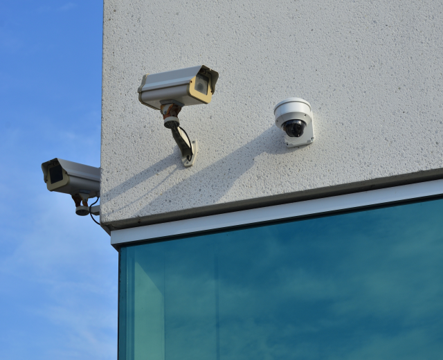 Zwei Überwachungskameras an der Seite eines Gebäudes montiert, mit einer Glaswand im Vordergrund und einem klaren blauen Himmel im Hintergrund.