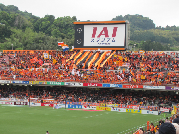 Ein Fußballspiel in einem Stadion mit einer großen Menge, einem grünen Rasen, einem Tor, Bannern, Fahnen, einem großen Bildschirm, Bäumen und einem klaren blauen Himmel.