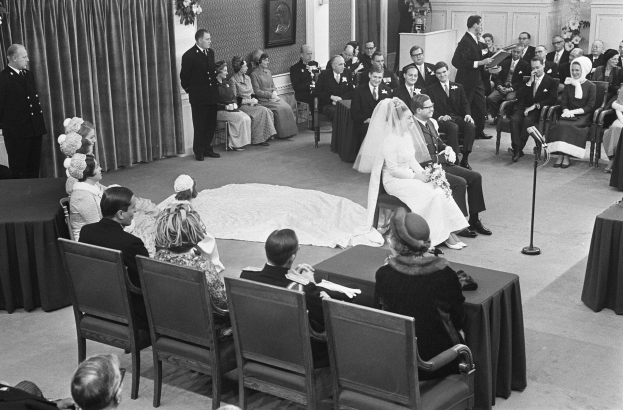 Schwarzes und weißes Foto einer Hochzeitszeremonie mit sitzenden Gästen, einem Mann mit Mikrofon, einer Frau mit Blumenstrauß, einem Podium mit Mikrofon, Blumenschmuck, einem Vorhang, einem gerahmten Foto an der Wand und einer Tür im Hintergrund.