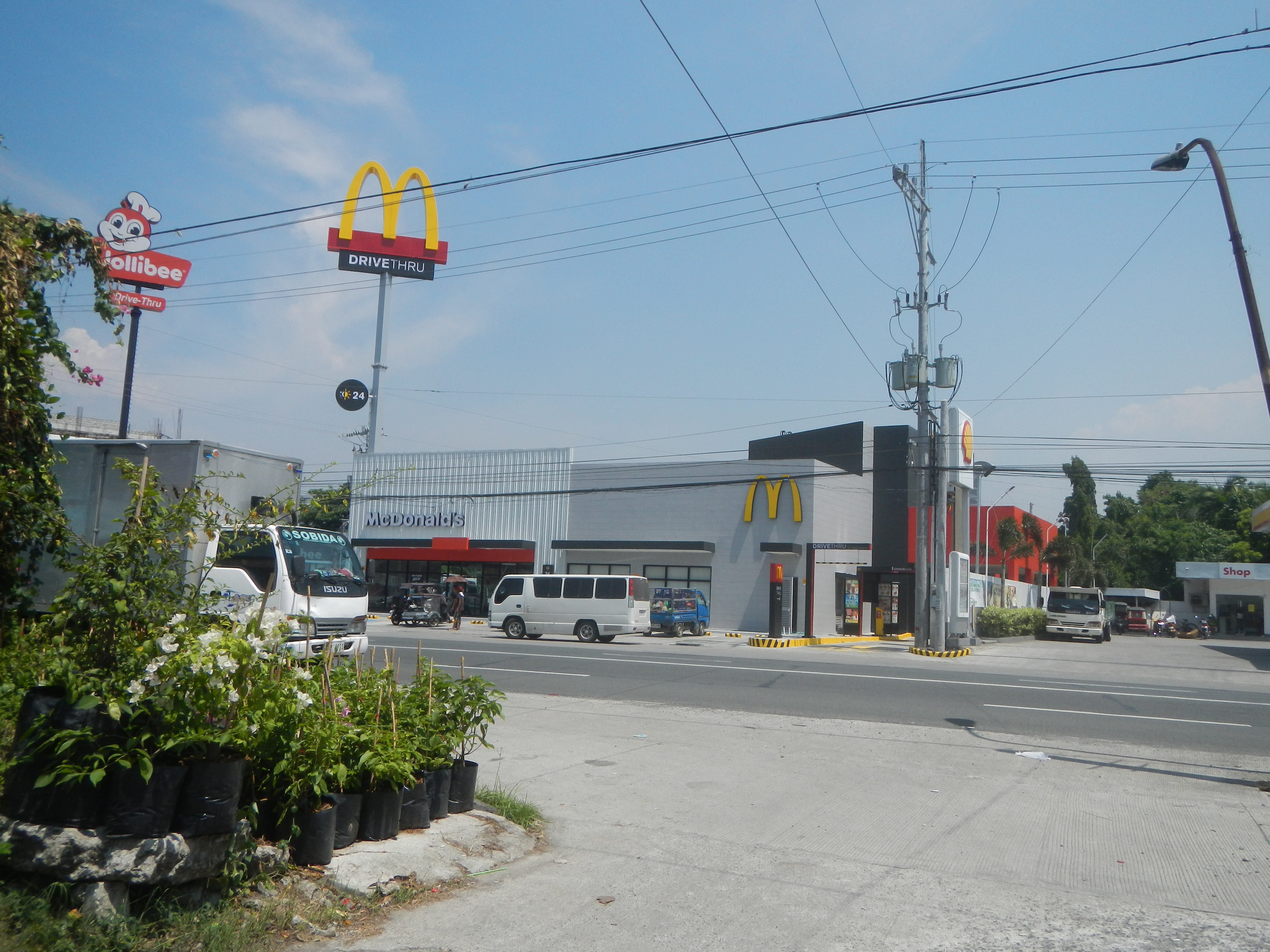 Ein McDonald's-Restaurant an einer Straßenecke mit Fahrzeugen, Gebäuden, Polen, Lampen, Schildern, Bäumen, Pflanzen, Gras im Vordergrund und einem bewölkten Himmel im Hintergrund.