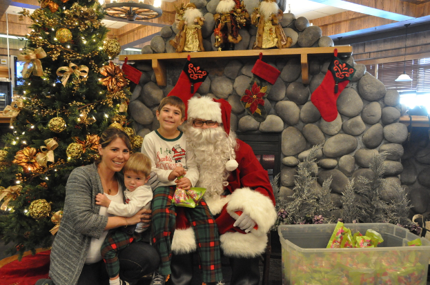 Eine Frau, Kinder und ein als Santa Claus verkleideter Mann mit Brille und Handschuhen stehen neben einem geschmückten Weihnachtsbaum, einer Wand mit Steinen, Spielzeugen, Décor und einem Korb mit Gegenständen, mit Lichtern im Hintergrund.