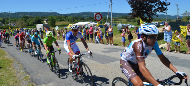 Gruppe von Radfahrern fährt an Zuschauern vorbei, einige tragen Helme und Schutzbrillen, andere halten Fahnen, mit Bäumen, Pfählen, Schildern, Fahrzeugen, Zelten, Gebäuden, Hügeln und einem klaren blauen Himmel im Hintergrund.