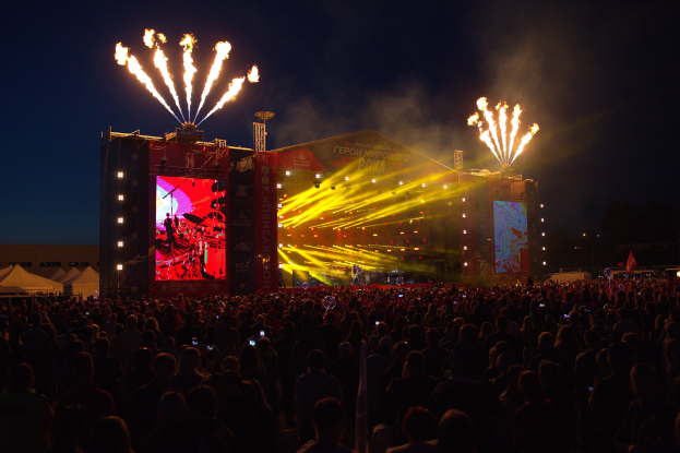 Eine große Menschenmenge auf einem Musikfestival schaut Performern auf einer hell erleuchteten Bühne zu, mit Zelten links und Feuerwerk in einem dunklen Himmel dahinter.