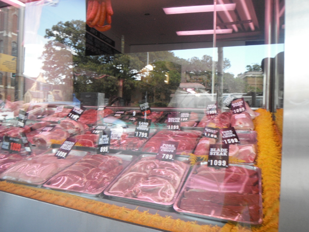 Eine Glasvitrine in einem Metzgerladen, die Tabletts mit Fleisch und Preisschildern zeigt, mit Gebäuden und Bäumen im Fenster und einer Decke oben im Bild.
