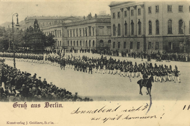 Ein Schwarz-Weiß-Foto eines Umzugs in Berlin, Deutschland, mit Menschen zu Fuß und zu Pferd, einer Statue, Straßenlaternen, Bäumen, Gebäuden mit Fenstern und einem bewölkten Himmel, mit Text am unteren Rand.