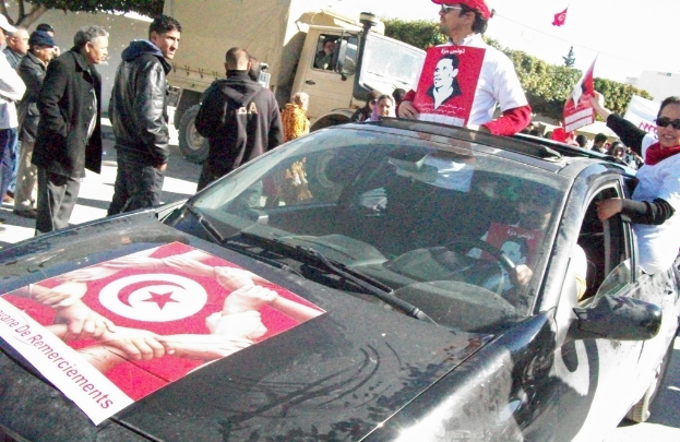 Drei Personen in einem Auto mit roten Plaketten auf ihren Hemden, umgeben von einer Gruppe von Menschen, mit einem Lastwagen dahinter.