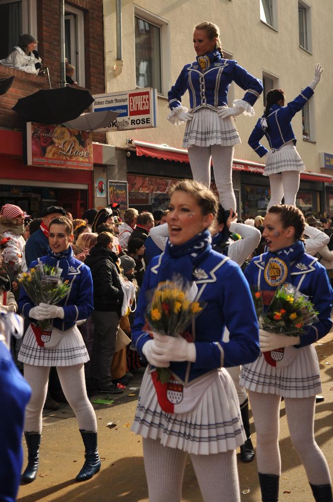 Eine Gruppe von Frauen, die Gymnastik auf einer Straße machen, während andere Blumensträuße halten und singen, und Zuschauer zusehen; im Hintergrund ist ein mehrstöckiges Gebäude mit Fenstern, Plakaten und Schildern zu sehen.