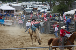 Zwei Menschen reiten auf Pferden bei einem Wettbewerb namens Charreada, umgeben von Gruppen von Menschen, Fahnen und Bannern, Zeltplanen, geparkten Fahrzeugen und Bäumen im Hintergrund.
