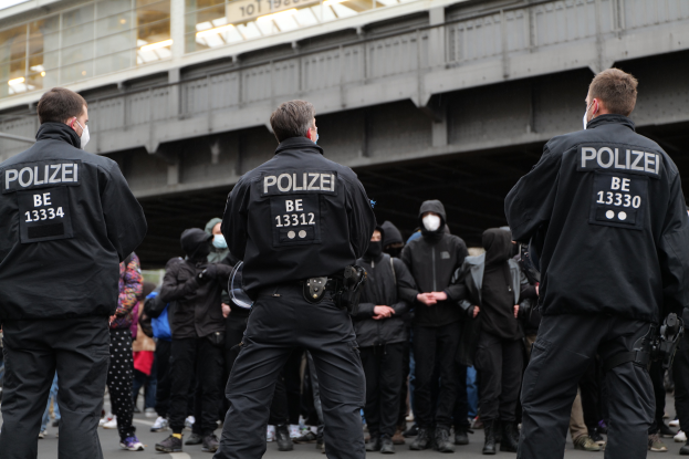Eine Gruppe uniformierter Polizisten steht vor einer Menge von Menschen in schwarzen Uniformen und Masken, im Hintergrund eine Stadtbrücke und ein Gebäude.