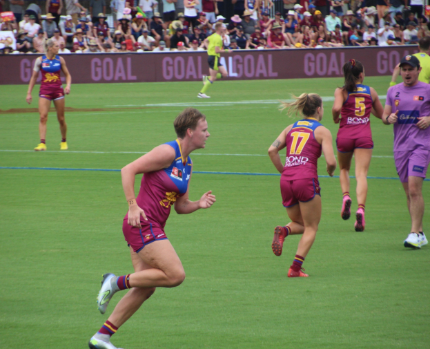 Frauen in Sportkleidung spielen Australian Rules Football auf einem Rasenfeld mit Zuschauern hinter Absperrungen im Hintergrund.
