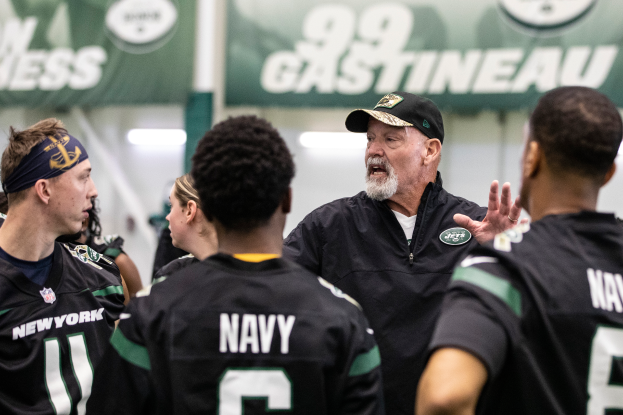 Eine Gruppe von Männern auf einem Feld, mit einem Mann in der Mitte mit einer Mütze, wahrscheinlich Jets Head Coach Mike McCarthy, der mit seinem Team spricht, mit Bannern und Lichtern im Hintergrund, die auf ein Stadion oder eine Sportanlage hindeuten.