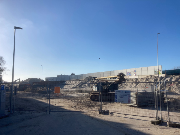 Eine Baustelle mit einem großen Haufen Erde in der Mitte, umgeben von einem Zaun, Pfählen, Lampen, Schildern und Bäumen, unter einem klaren Himmel, Teil einer neuen Einkaufszentrumsbau in den Niederlanden.