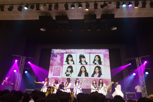 Eine Gruppe von Menschen performs auf einer hell erleuchteten Bühne mit Trägern, während ein Sänger ein Mikrofon hält und ein Musiker ein Instrument spielt, während die Menge darunter das Ereignis aufzeichnet und ein großer Bildschirm Bilder der K-Pop-Gruppe BK48 anzeigt.