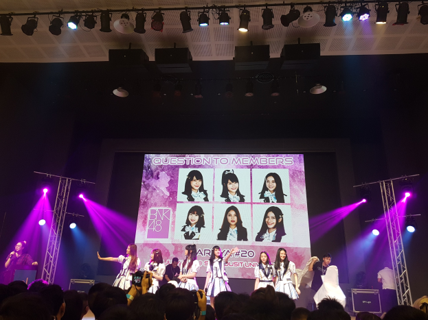 Eine Gruppe von Menschen performs auf einer hell erleuchteten Bühne mit Trägern, während ein Sänger ein Mikrofon hält und ein Musiker ein Instrument spielt, während die Menge darunter das Ereignis aufzeichnet und ein großer Bildschirm Bilder der K-Pop-Gruppe BK48 anzeigt.