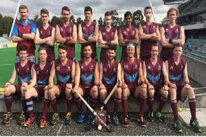 Eine Gruppe von Männern, die auf einem Feld mit Hockey-Schlägern feiern, mit Bäumen und einem Stadion mit Zuschauern im Hintergrund.