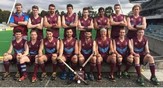 Eine Gruppe von Männern, die auf einem Feld mit Hockey-Schlägern feiern, mit Bäumen und einem Stadion mit Zuschauern im Hintergrund.