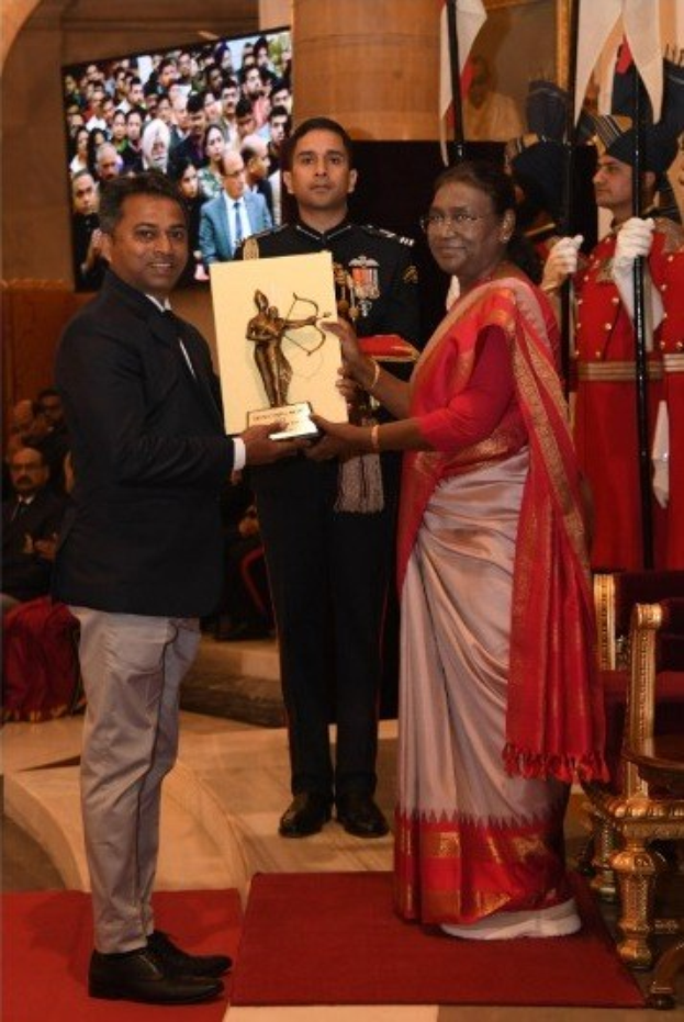 Ein Mann in einem schwarzen Blazer, weißem Hemd und grauen Hosen hält eine Trophäe, während er neben einer Frau in einem aschfarbenen Sari auf einem roten Teppich an einer Preisverleihung steht, mit Menschen, Stühlen, Fahnen und einem Bildschirm im Hintergrund.