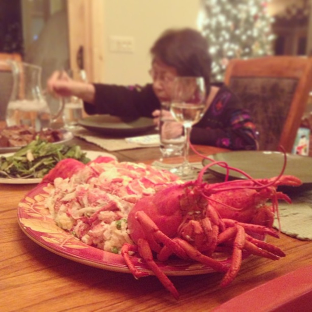 Eine Frau sitzt an einem Tisch und hält Stäbchen, mit Essen, einem Wasserglas und anderen Gegenständen auf dem Tisch, einem Weihnachtsbaum im oberen rechten Eck und einer Tür mit Steckdosen im Hintergrund.