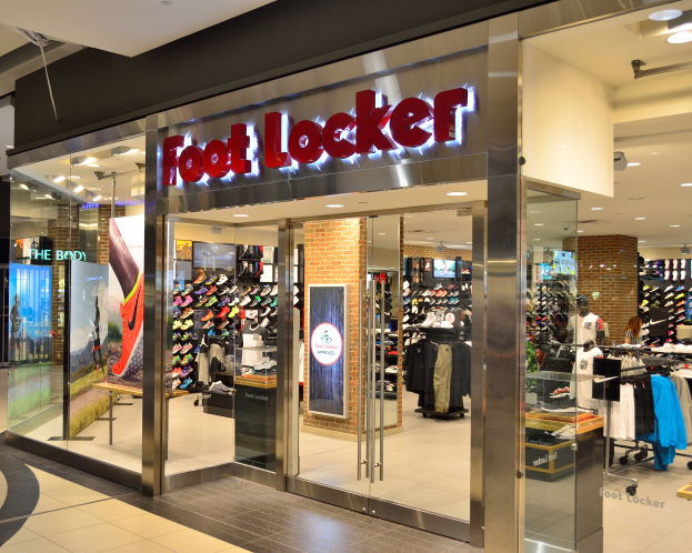 Ein Foot Locker Geschäft in einem Einkaufszentrum mit Glaswänden und -türen, Kleidung an Bügeln, bekleidete Schaufensterpuppen, Texttafeln, Deckenleuchten und verschiedene andere Gegenstände.