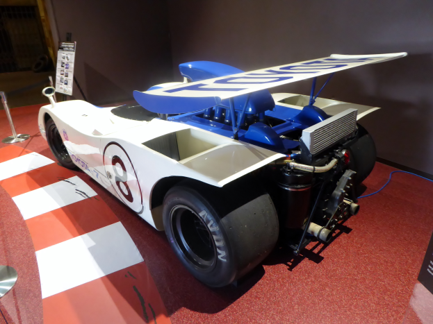 Ein weiß-blauer Rennwagen in einem Museum ausgestellt, umgeben von Absperrungen und Schautafeln, mit einer Wand im Hintergrund, die auf das Goodwood Festival of Speed hinweist.