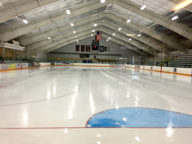 Große Indoor-Eisfläche mit Deckenbeleuchtung, Werbetafeln, einer Rückwand, Treppen mit Geländern und einigen Menschen, die einem Hockeyplatz ähneln.