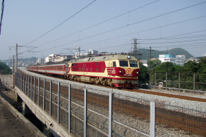 Ein roter und gelber Zug fährt auf Schienen, mit einer Brücke auf der einen Seite und einem Geländer auf der anderen, während Strommasten, Bäume, Gebäude und ein klarer blauer Himmel im Hintergrund zu sehen sind.