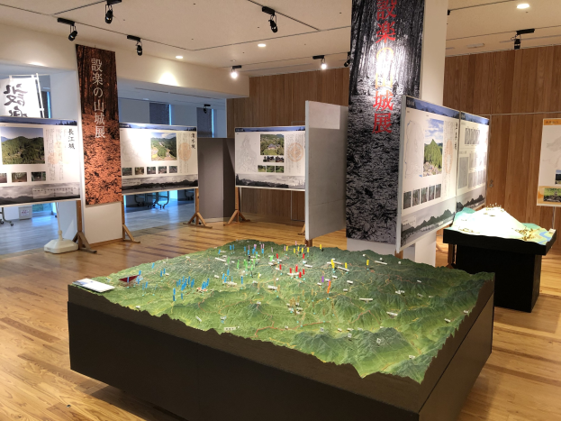 Ausstellungsraum mit verschiedenen historischen Karten Japans, einschließlich montierter Anzeigen, Banner und einem zentralen Tisch mit einer Karte darunter, unter Deckenleuchten an einer hölzernen Wand.