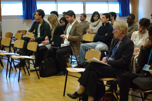 Mehrere Personen sitzen in einem Raum auf Stühlen und tragen ID-Karten, wobei ein Mann einen Laptop und eine Frau einen Stift hält, umgeben von leeren Stühlen, Fenstern mit Vorhängen und einer Tasche auf dem Boden.