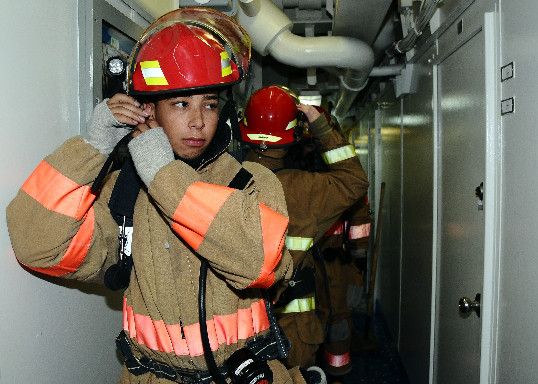 Feuerwehrleute in Uniform während eines Trainings in einem Raum, mit Rohren und Equipment im Hintergrund.