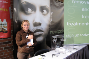 Eine Frau steht lachend und hält ein Blatt Papier in der Hand, mit einem Stuhl, einem mit einer Decke bedeckten Tisch, einer Glasbox voller Papiere, Stiften, einem Handy und anderen Gegenständen, sowie zwei Bannern und einer Wand im Hintergrund.