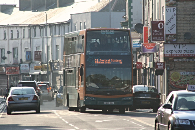 Eine Straße mit Autos und einem Bus im Vordergrund, im Hintergrund Gebäude mit Wänden, Fenstern, Tellern und Dächern sowie Plakate und Banner an den Wänden und einem Laternenmast.