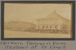 Eine Gruppe deutscher Gefangener aus Messines steht vor einem Gebäude unter einem klaren blauen Himmel, mit Text unten im Bild.
