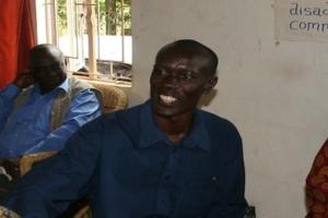 Menschen auf Stühlen sitzend mit einer Wand, einem Fenster und einem Vorhang im Hintergrund und Text in der oberen rechten Ecke.