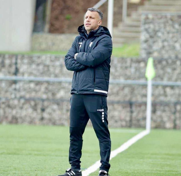 Ein Mann steht auf einem Fußballfeld mit verschränkten Armen, trägt eine schwarze Jacke und Hose, mit einem Zaun, Treppe und einer Wand im Hintergrund.