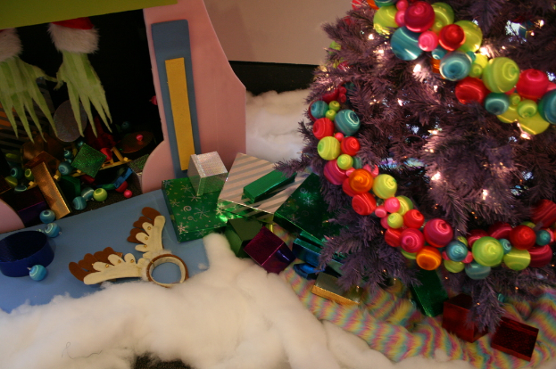 Eine Indoor-Szene mit einer Wand im Hintergrund, einem Baumwoll- und Wolltuch unten, einem geschmückten Weihnachtsbaum rechts, Geschenkboxen und dekorativen Gegenständen in der Mitte und einer Hütte mit Dekorationen und Baumwolle oben links.