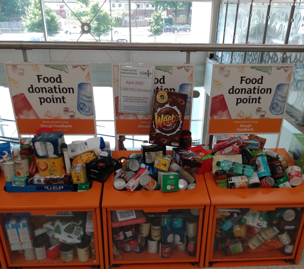 Orange Schübe mit Lebensmitteln wie Flaschen, Dosen und Kisten, neben Texttafeln, durch Glasfenster mit Fahrzeugen, Bäumen, Gebäuden und Himmel.