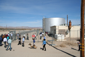 Eine spärlich besiedelte Landschaft mit wenigen Menschen, einem Hund, Häusern, einem Zaun und Strommasten unter einem klaren Himmel.